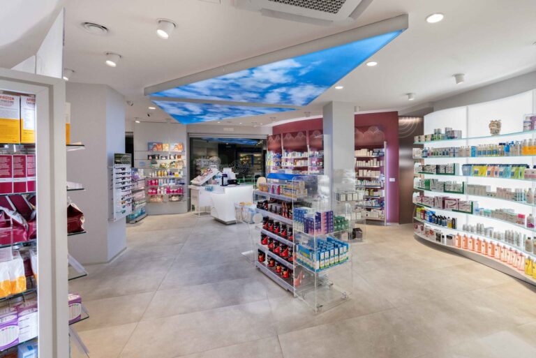 Panoramica farmacia con layout aperto, espositori centrali trasparenti e pareti illuminate per categorie prodotto