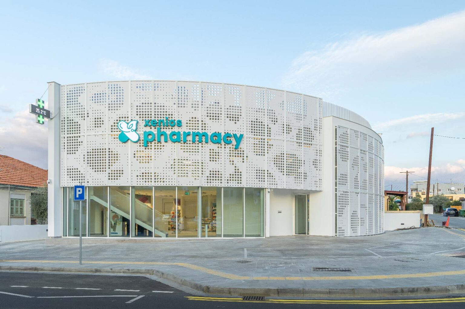 Vista esterna diurna della Xenios Xenofontos Pharmacy con facciata bianca traforata, vetrate al piano terra e parcheggio.